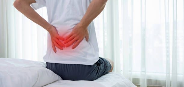 A man is kneeling on a bed holding his back in pain.