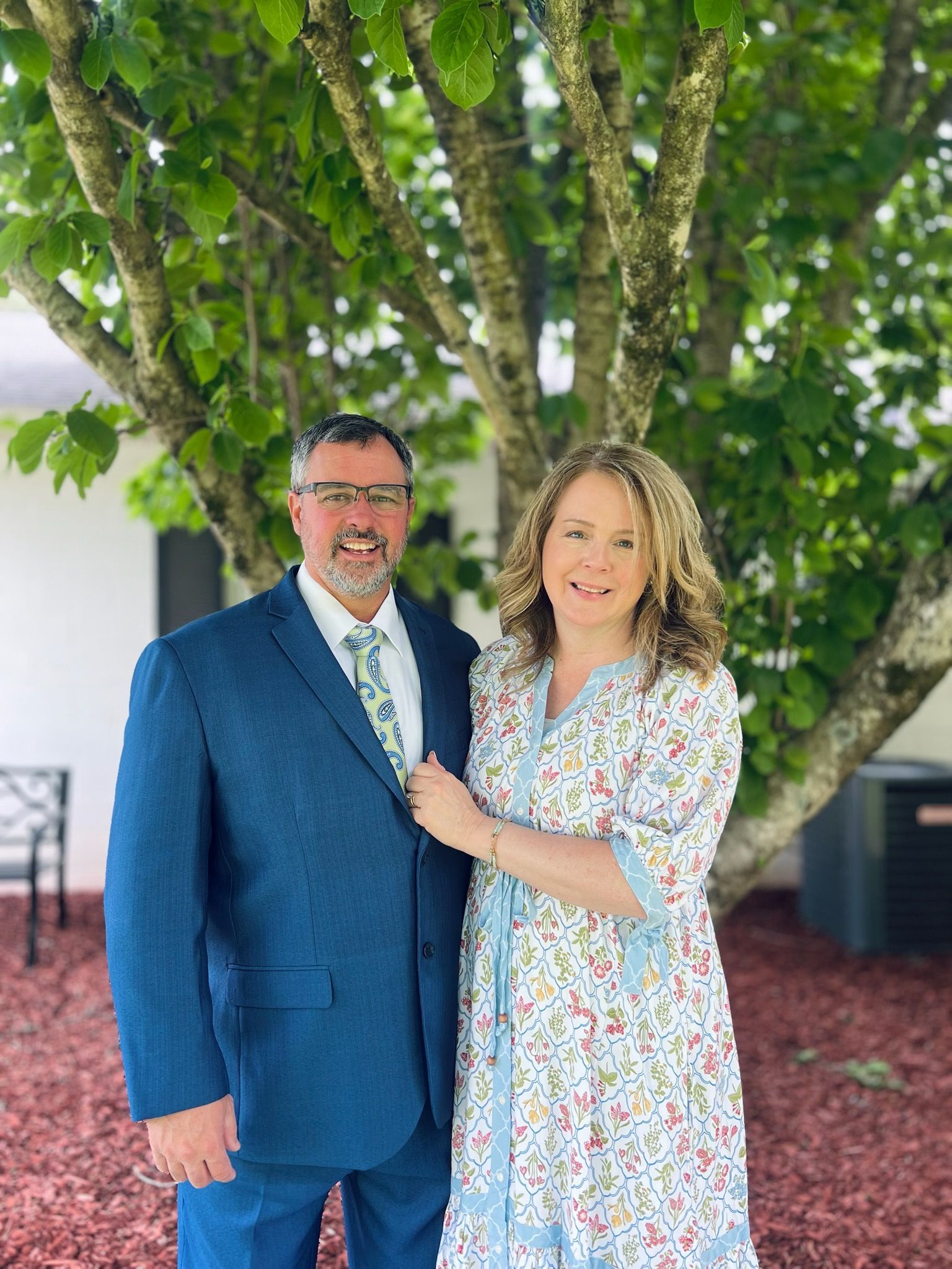 A man and woman stand together under a tree. The man wears a blue suit, and the woman wears a floral dress. They are smiling.
