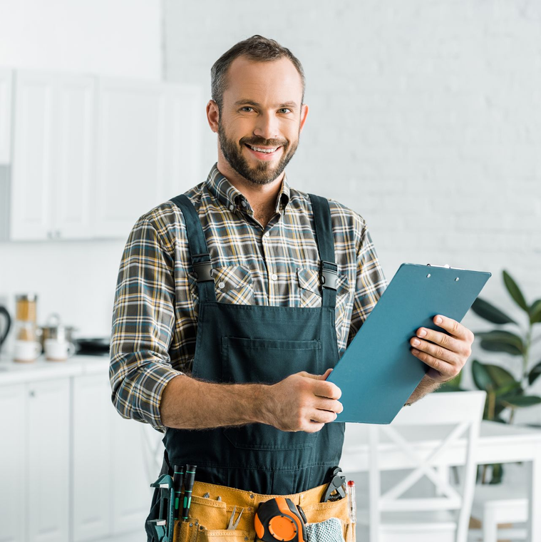 Smiling carpenter in apron holding a blue clipboard in a bright workshop