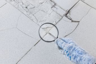 Cracked white tile floor under a magnifying glass, with a gloved hand inspecting damage