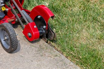 Red lawn edger cutting along a sidewalk beside grass, with part of a wheeled machine visible.