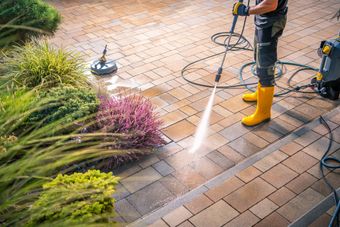 Worker pressure-washing a brick patio beside green and pink landscaping, wearing yellow boots