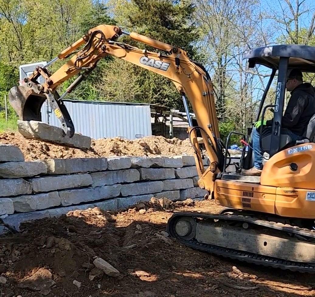 A man is driving an excavator on a construction site setting limestone blocks for retaining wall