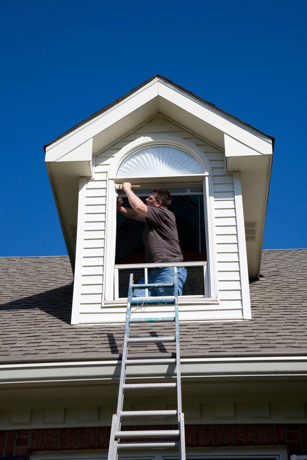 Person on ladder installing something in an arched window on a house under a clear, blue sky.