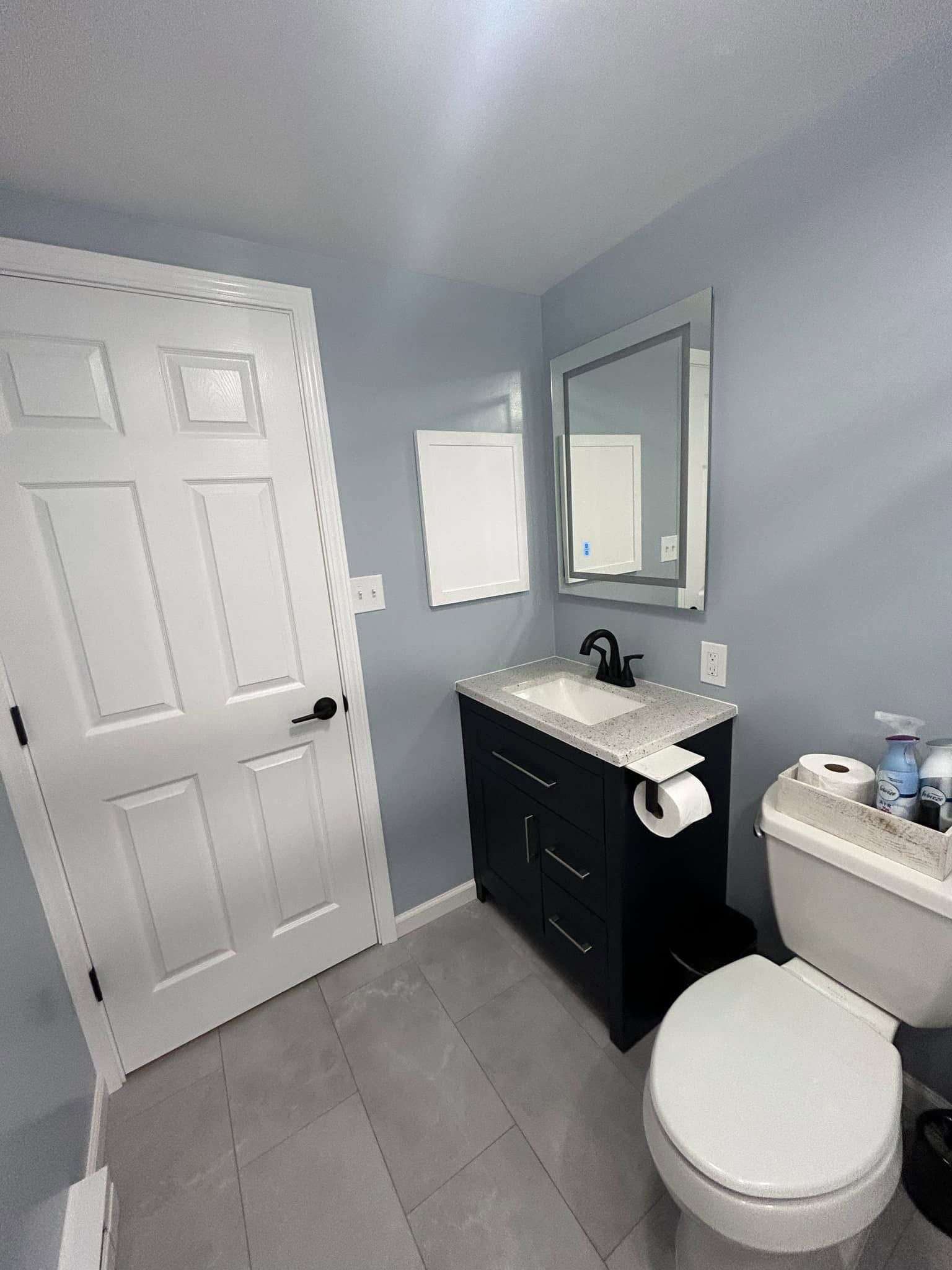 Small bathroom with gray tile floor, white door, black vanity, mirror, and toilet.