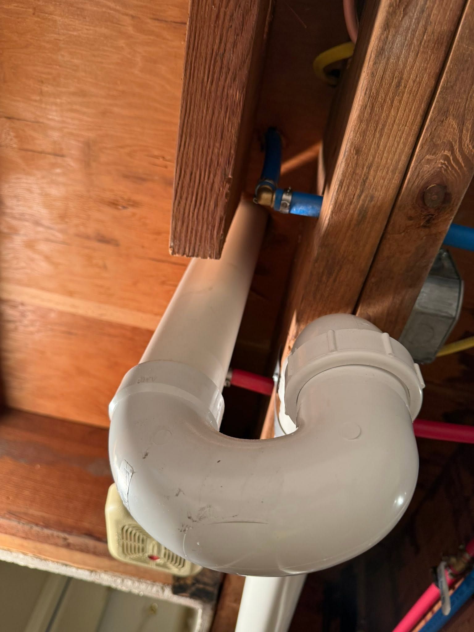 White PVC drain pipe elbow under wooden floor joists with a blue shutoff valve above it