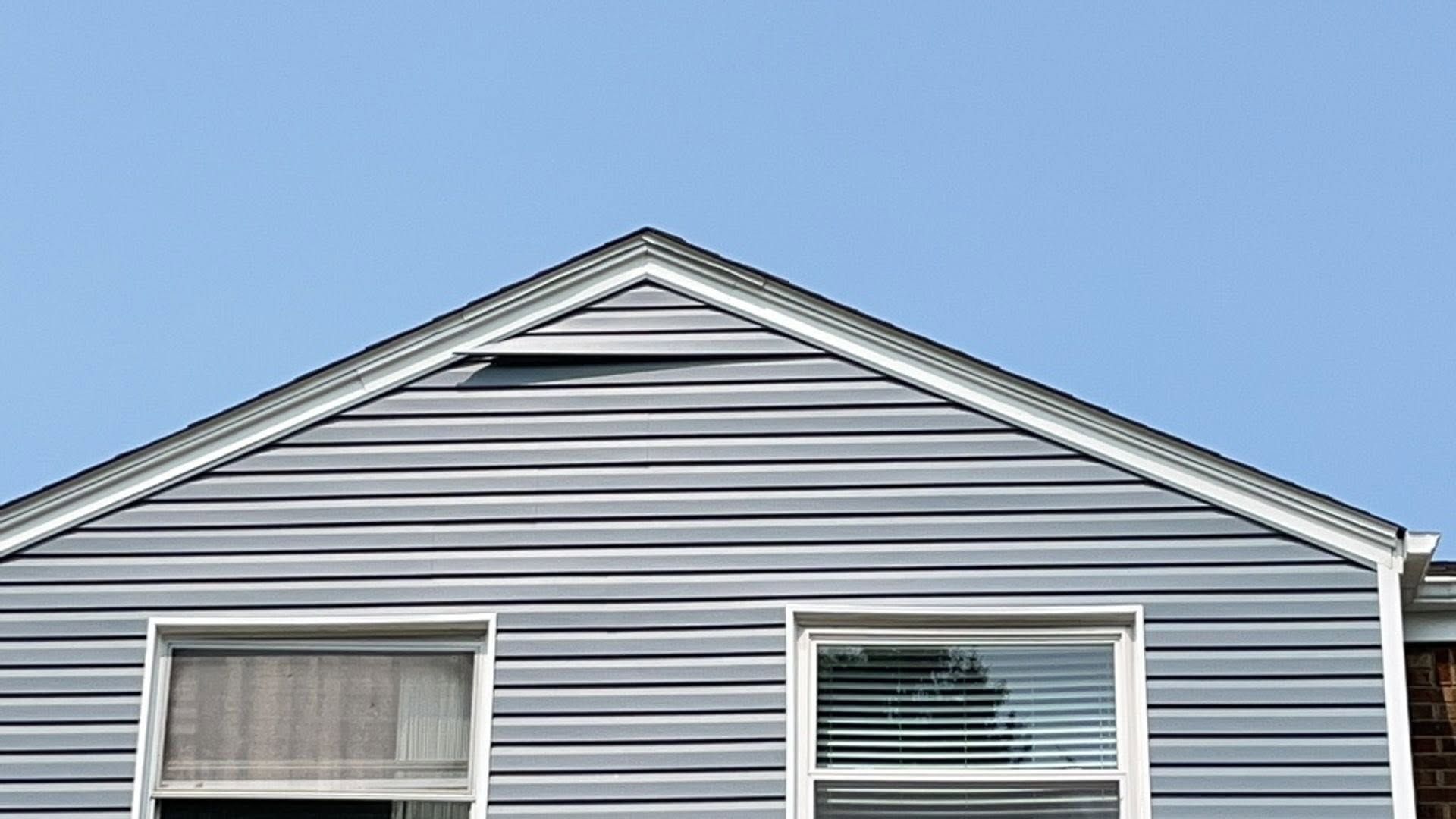 Blue house siding with white trim and two windows under a peaked roofline