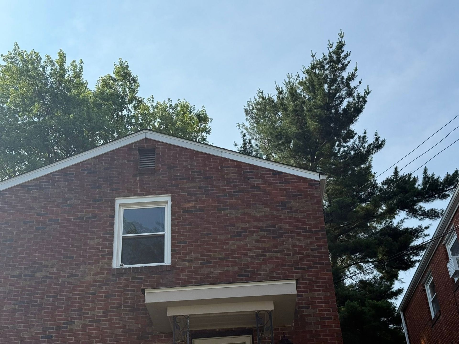 Red brick house with a white-trimmed window and awning under a blue sky, with trees behind it