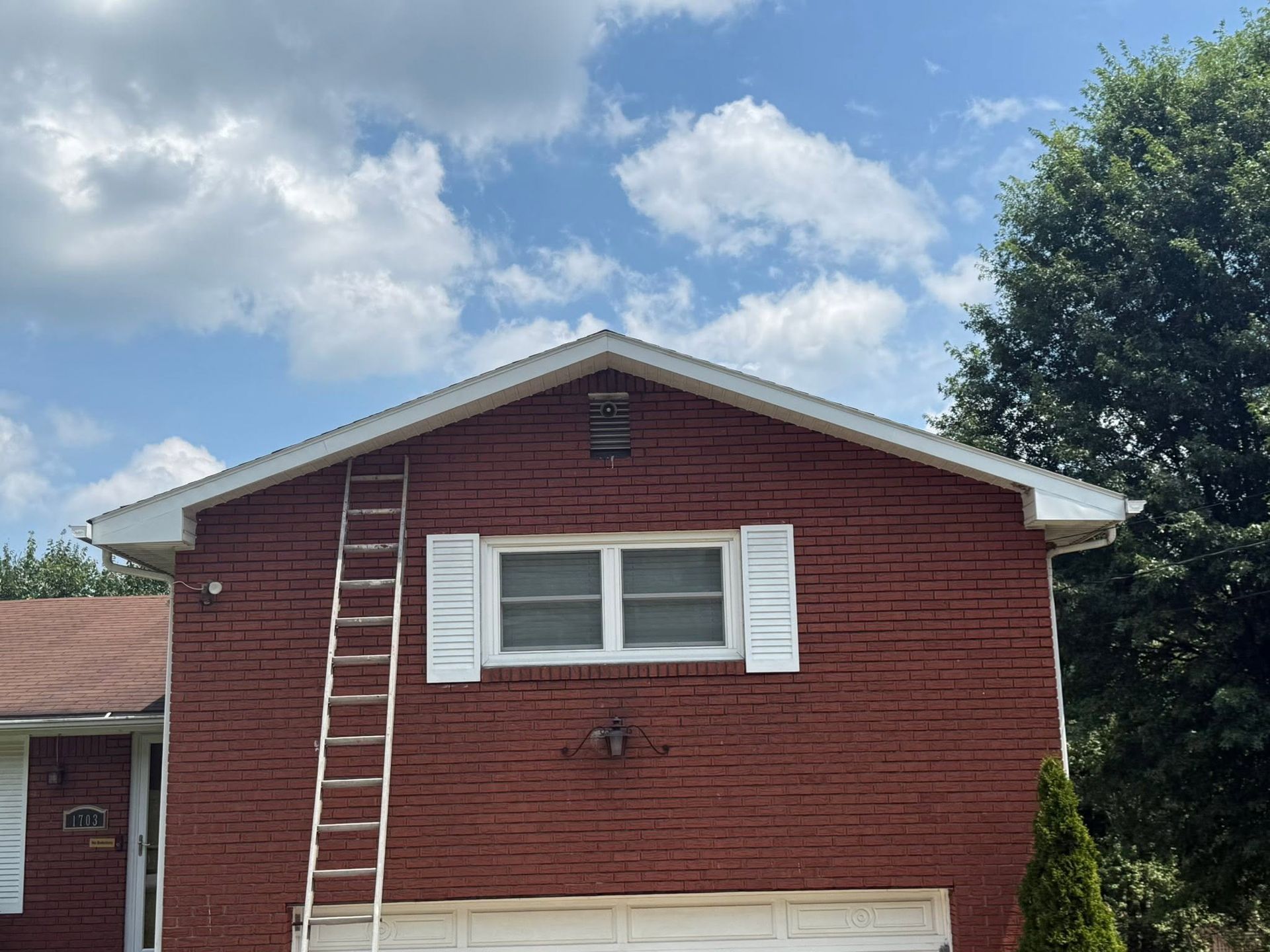 Red brick house with white shutters and a ladder on the roof under a blue, cloudy sky