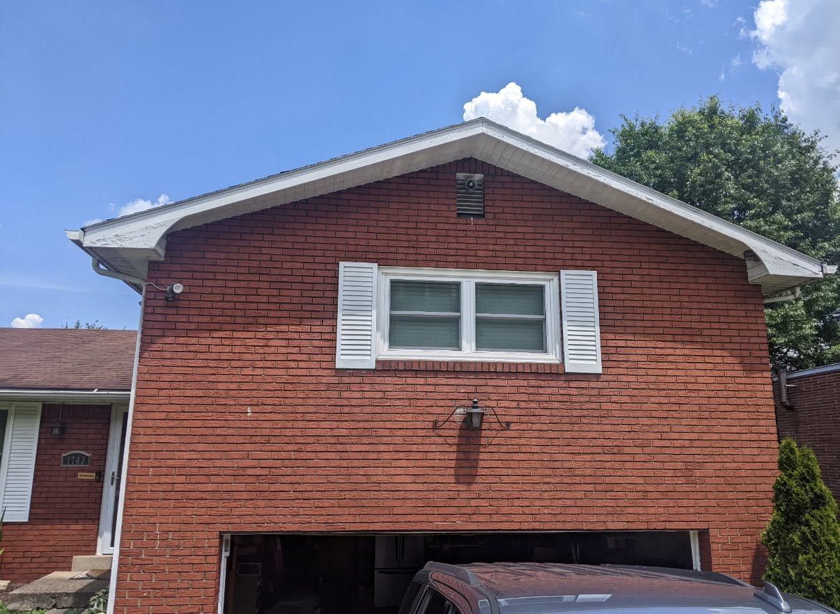 Red brick house exterior with white shutters and a garage under a blue sky.