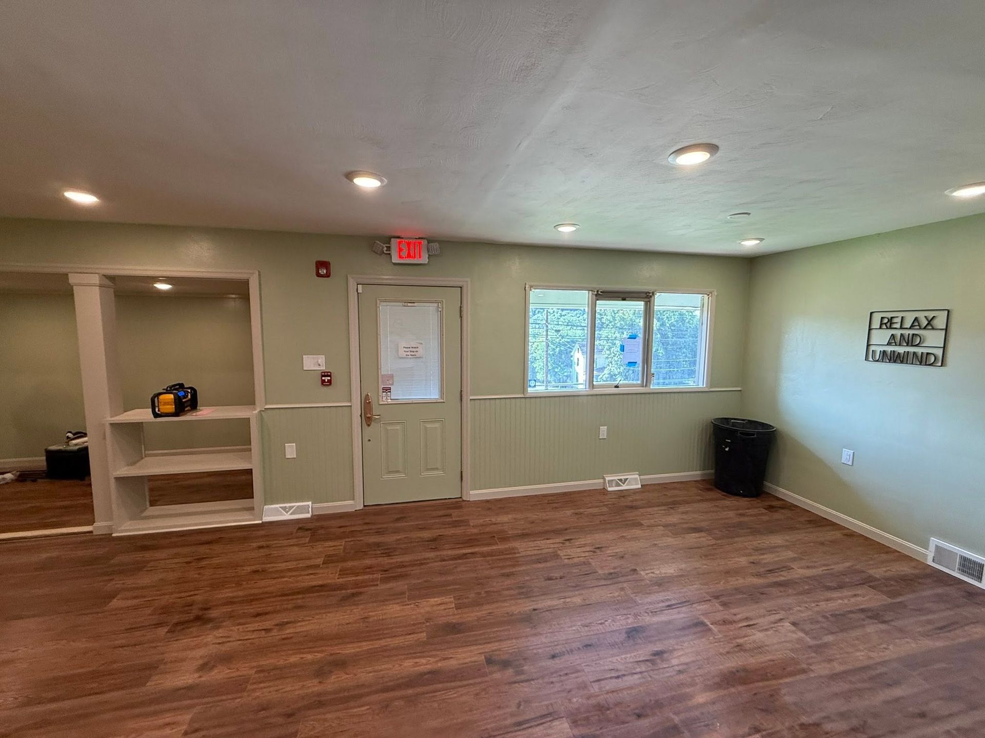 Empty room with wood floor, white walls, windows, and a door with exit sign.
