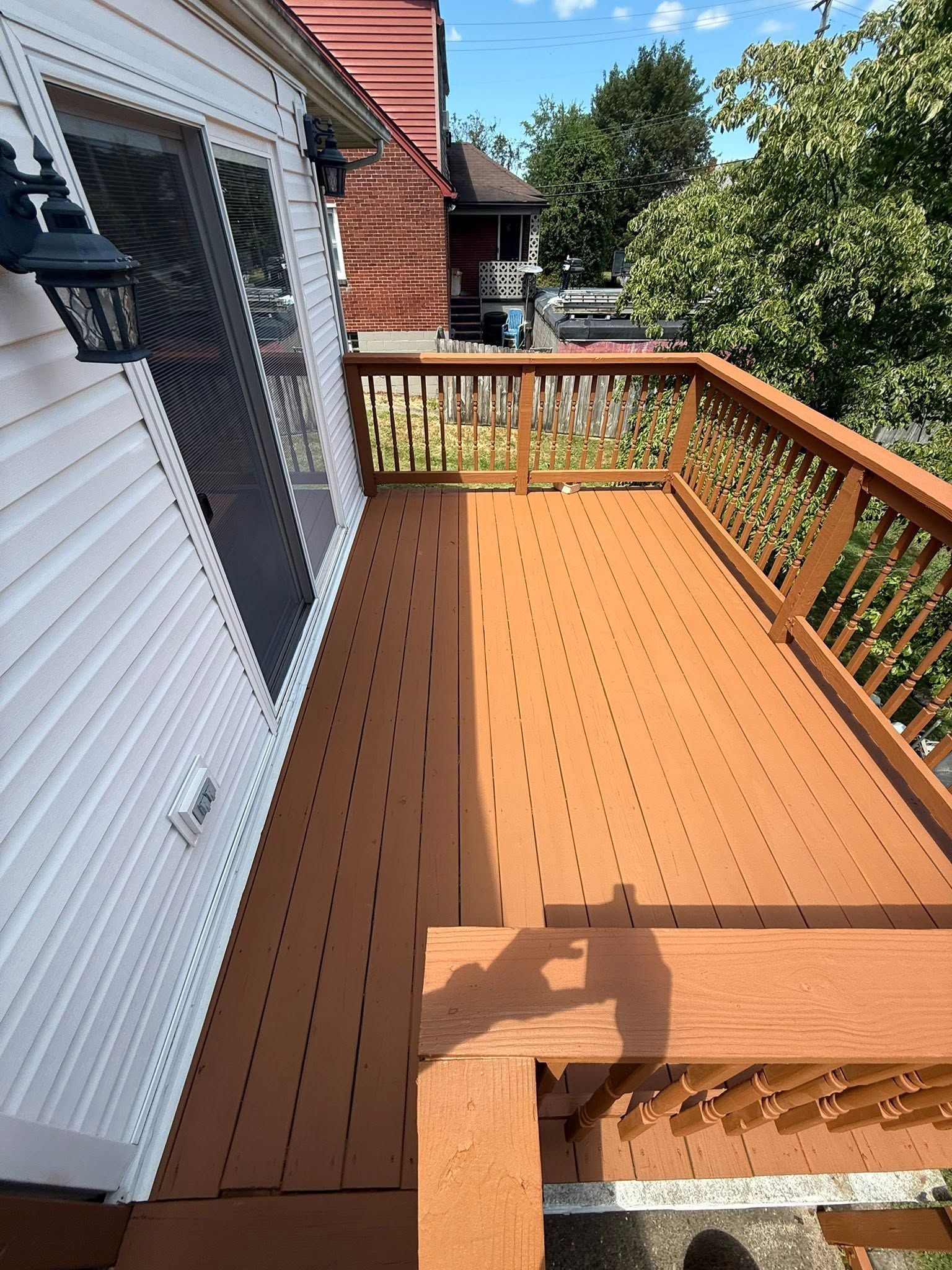 Wooden deck with orange-brown boards beside a white house, bordered by railing and trees beyond.