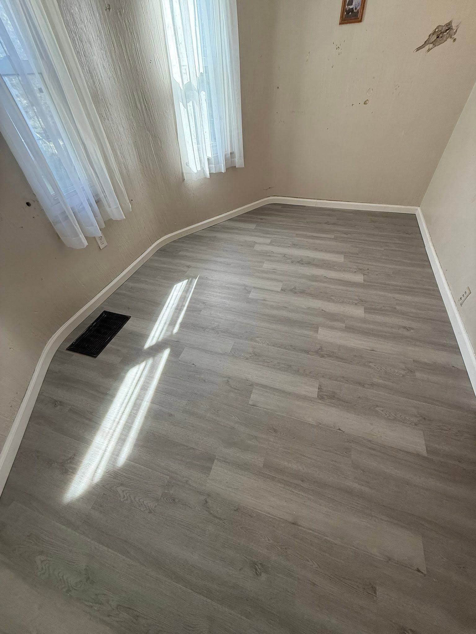 Empty room with light wood flooring, white curtains, and sunlight streaming across the floor.