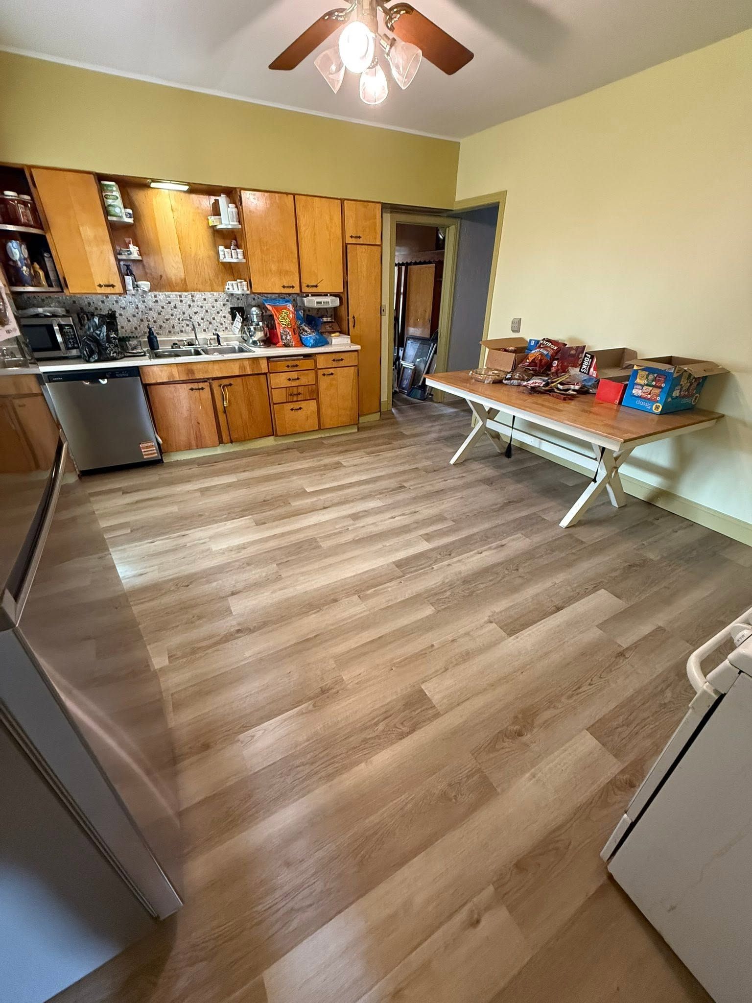 Small kitchen with wood cabinets, ceiling fan, wood-look floor, and a folding table with items on the right.