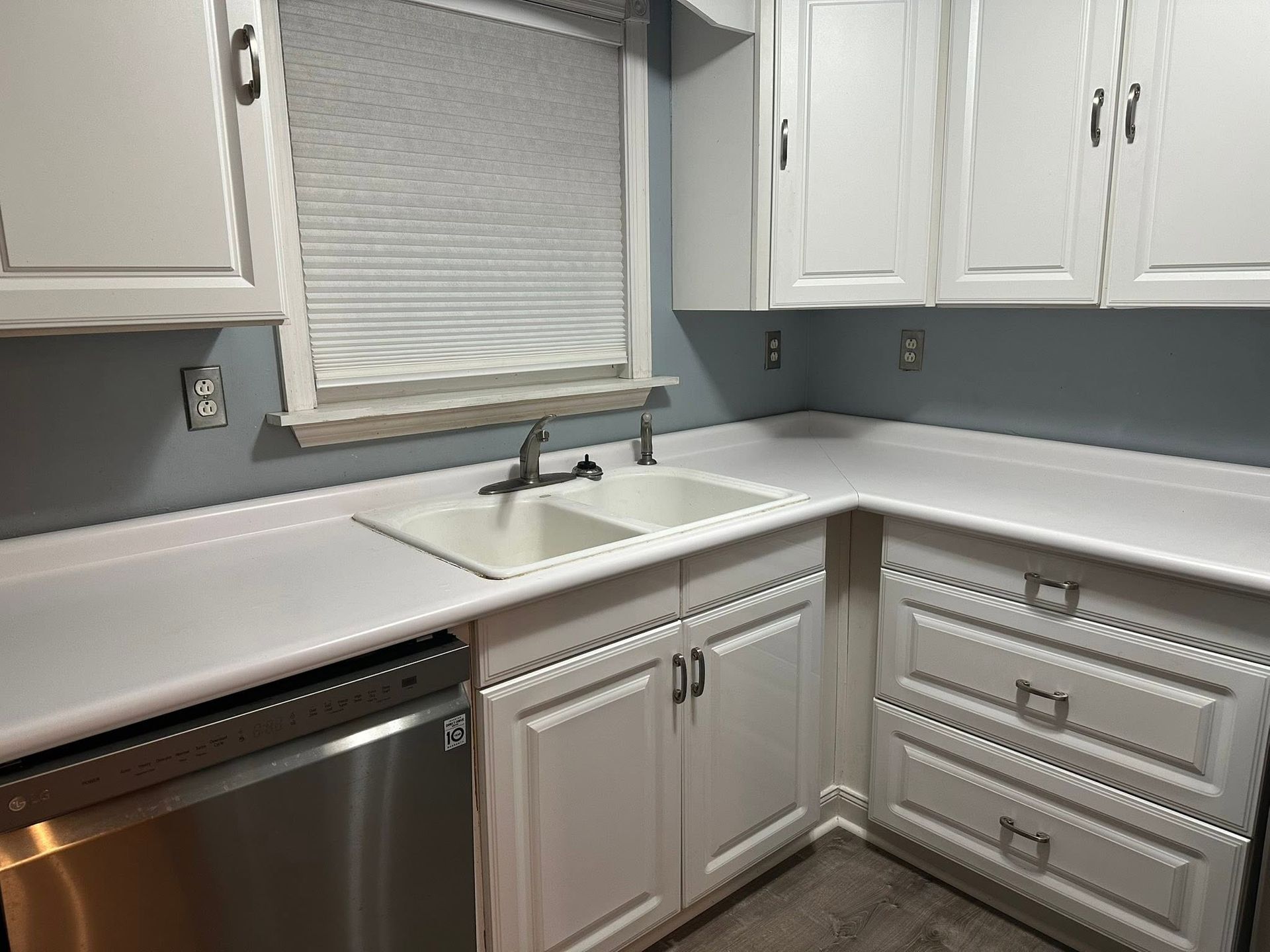 White kitchen corner with sink, window, cabinets, and stainless dishwasher