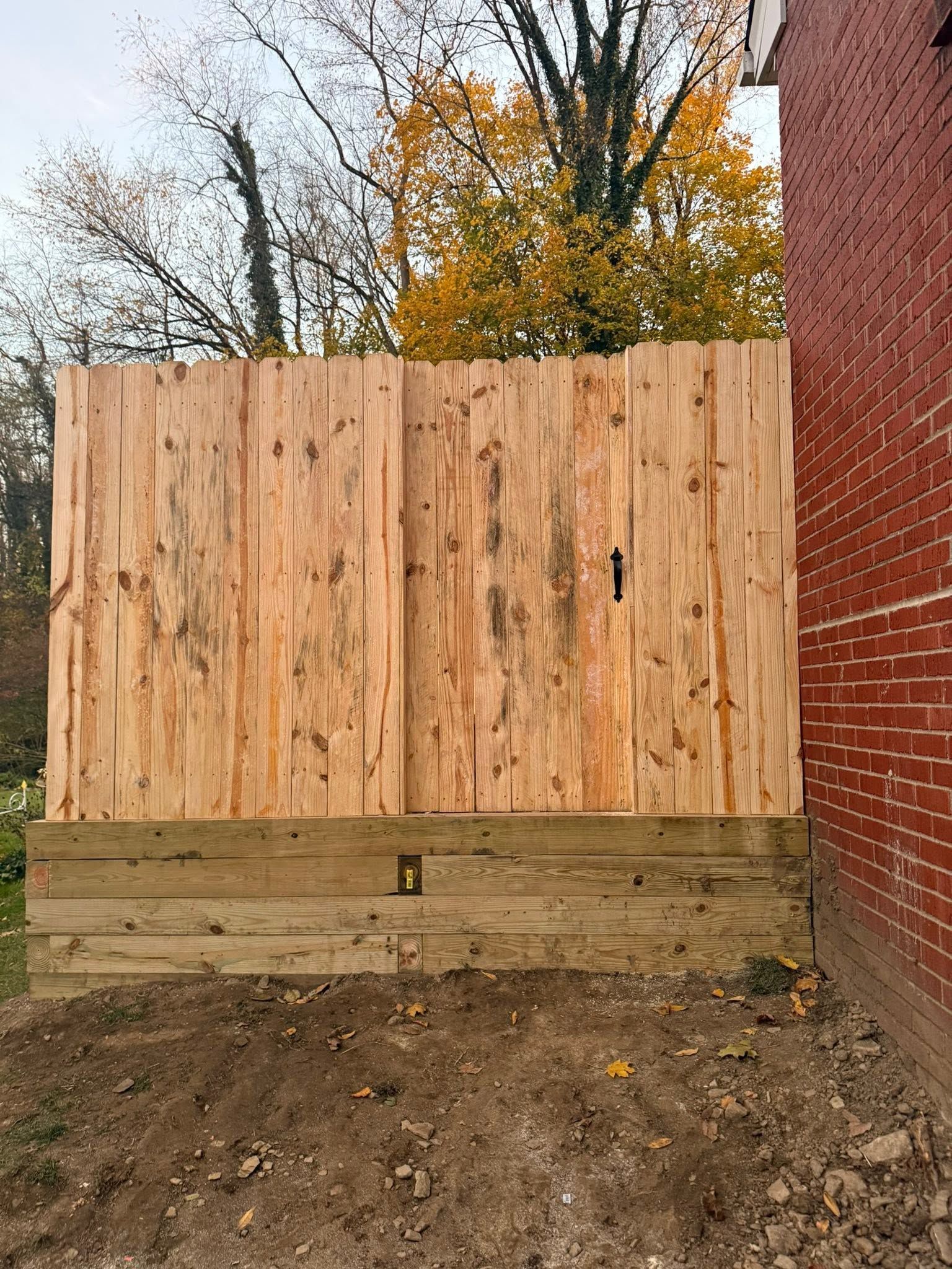 Wooden fence panel beside a red brick wall in a dirt yard, with autumn trees in the background