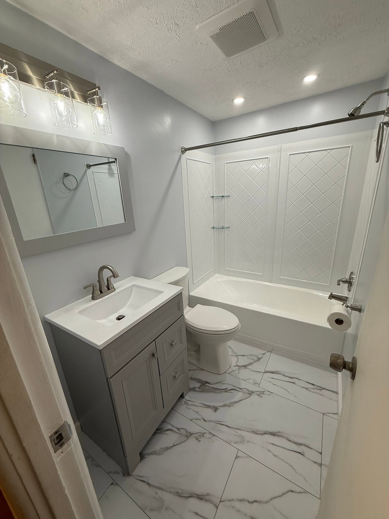 Small white bathroom with vanity, toilet, tub-shower combo, and marble-patterned floor and walls.