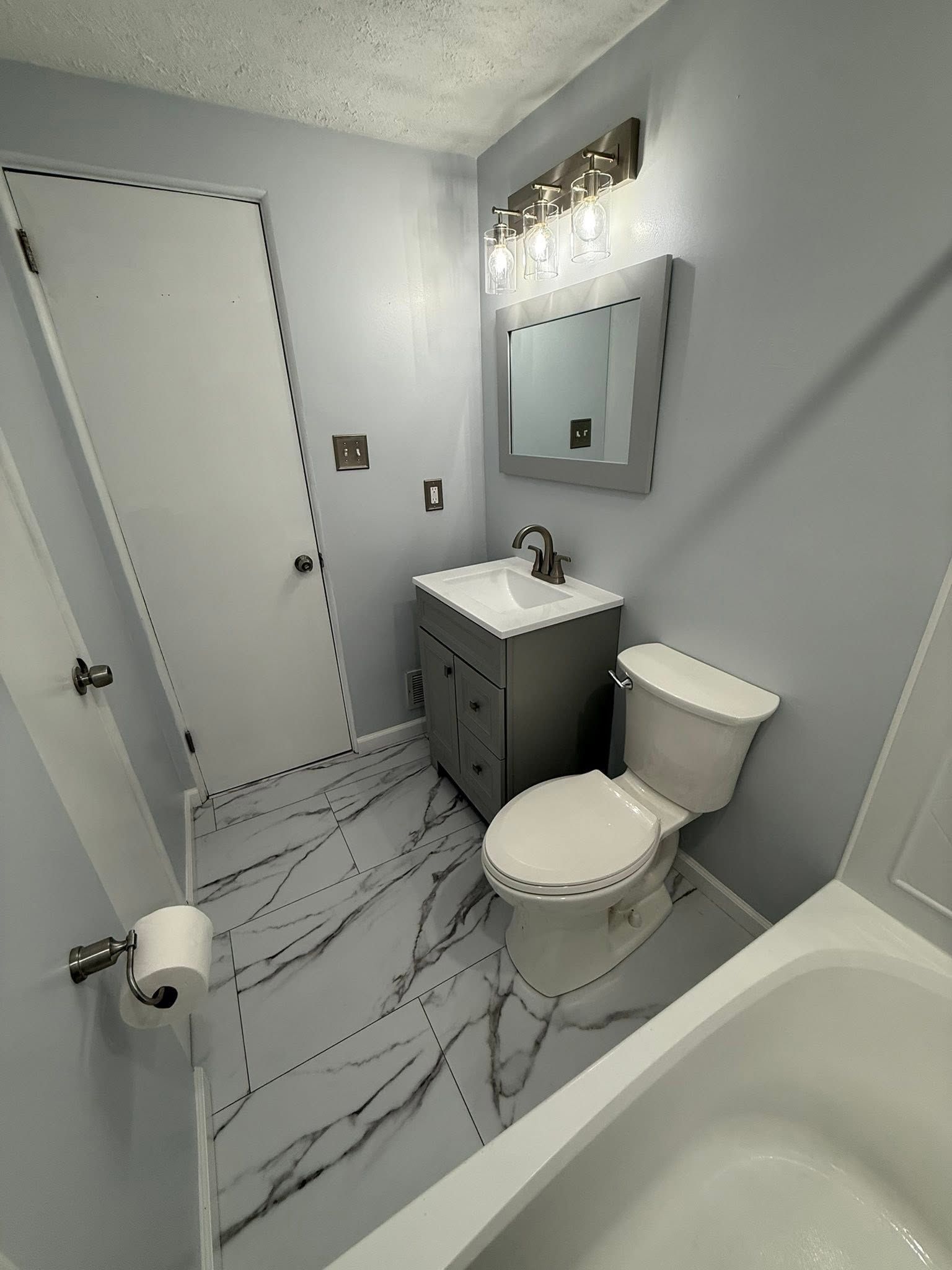 Small gray bathroom with bathtub, toilet, sink vanity, mirror, and marble-look floor tiles.
