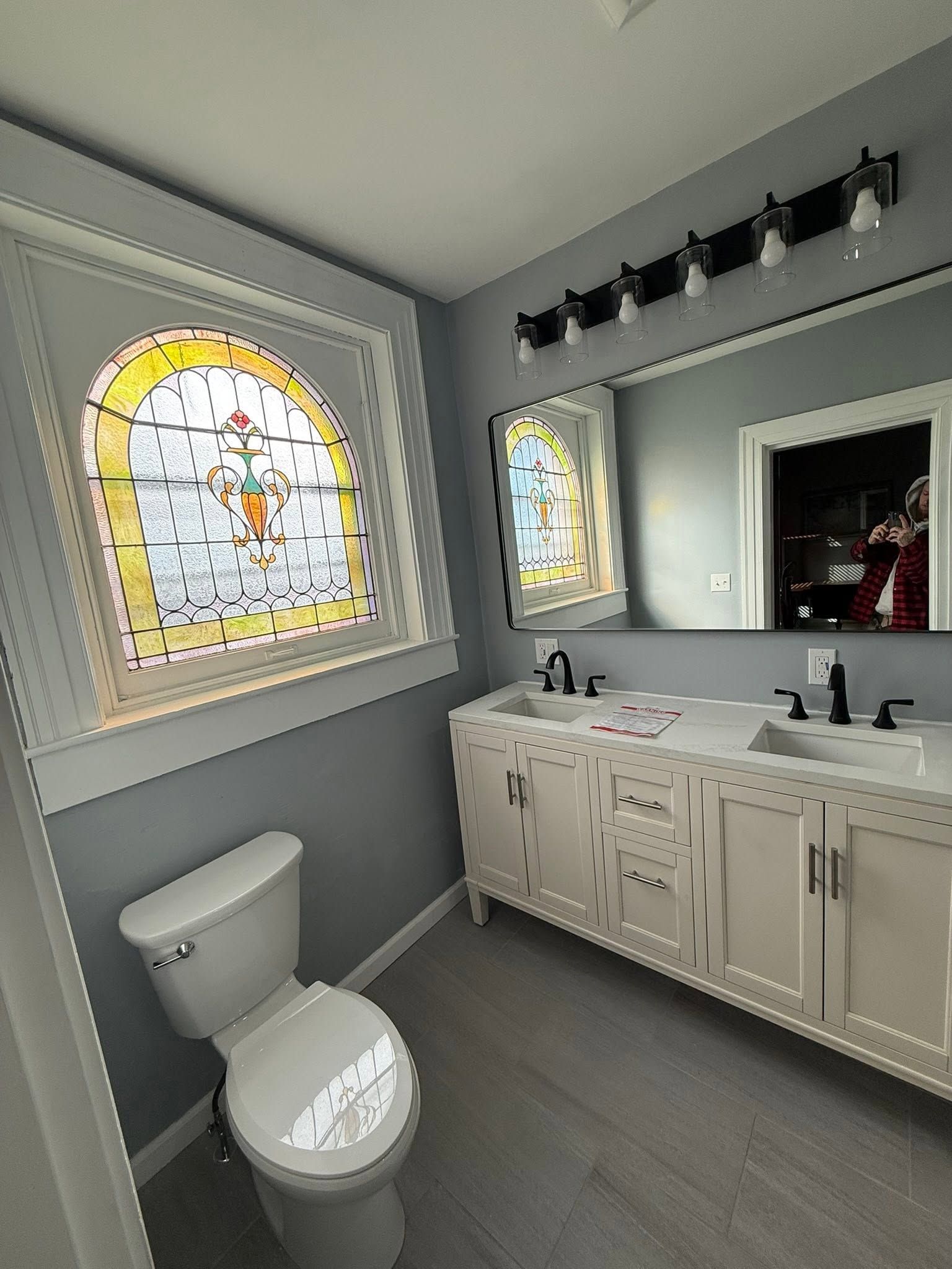 Bright gray bathroom with toilet, vanity, mirror, and stained-glass windows