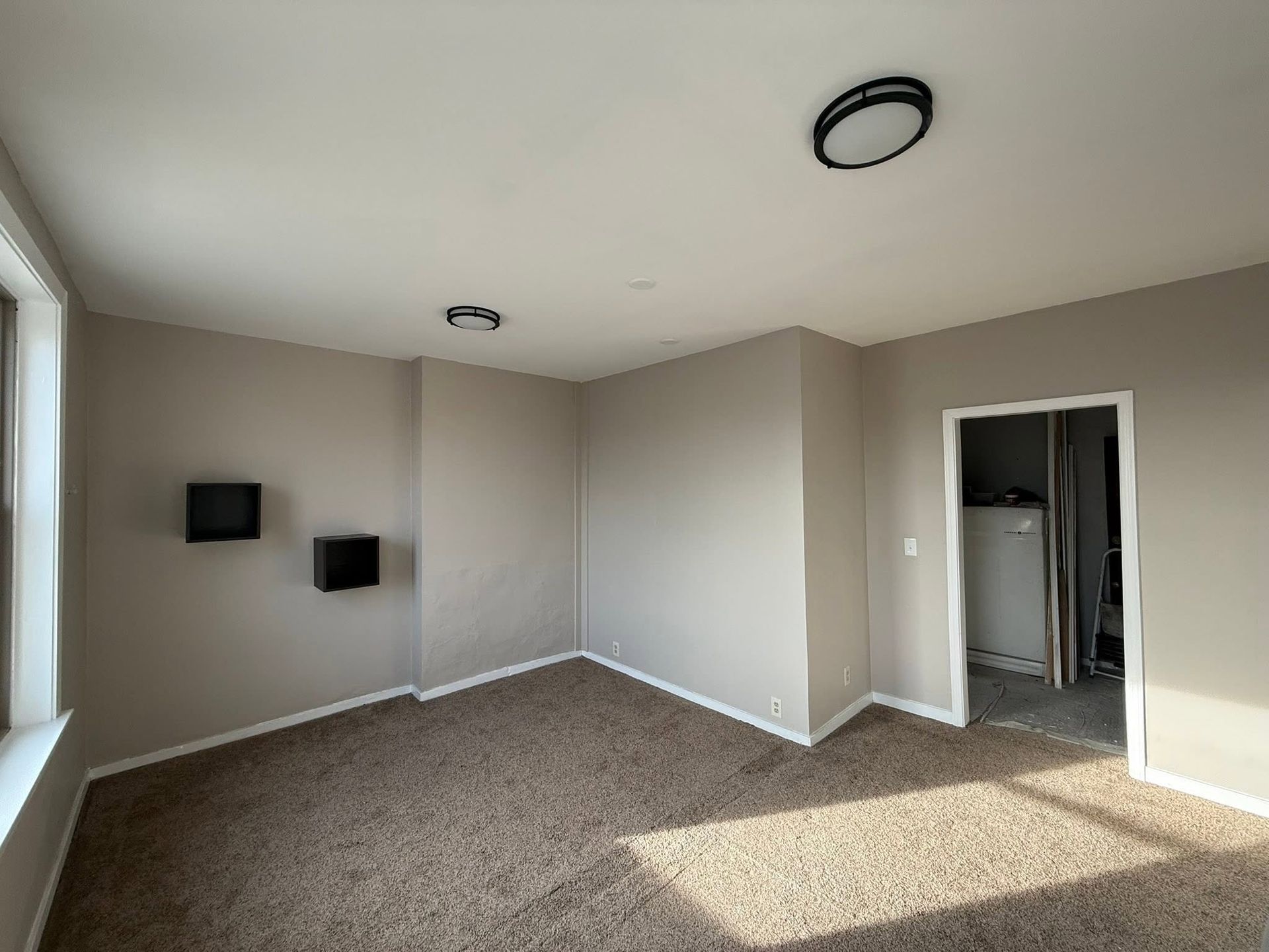 Empty beige carpeted room with wall-mounted TVs, ceiling light, and open doorway to a closet or bathroom