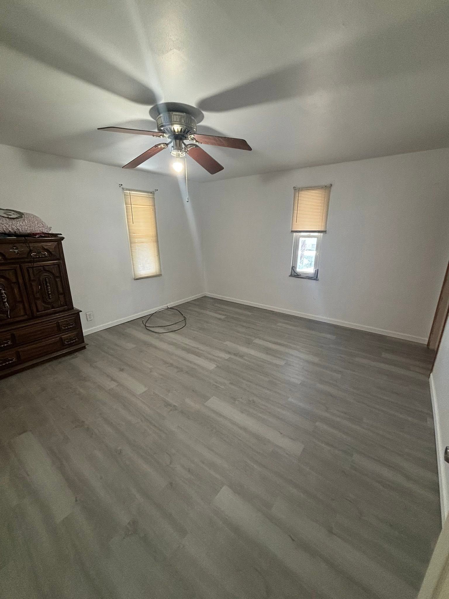 Empty bedroom with gray wood floors, white walls, two small windows, and a ceiling fan.