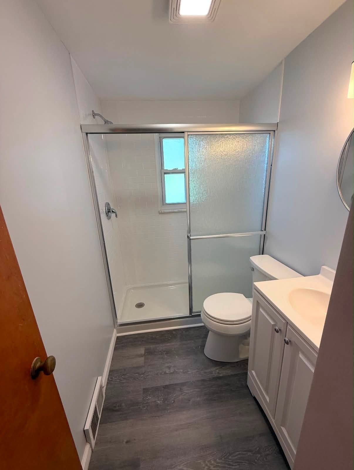 Narrow bathroom with walk-in shower, toilet, and vanity; gray floor and white walls.