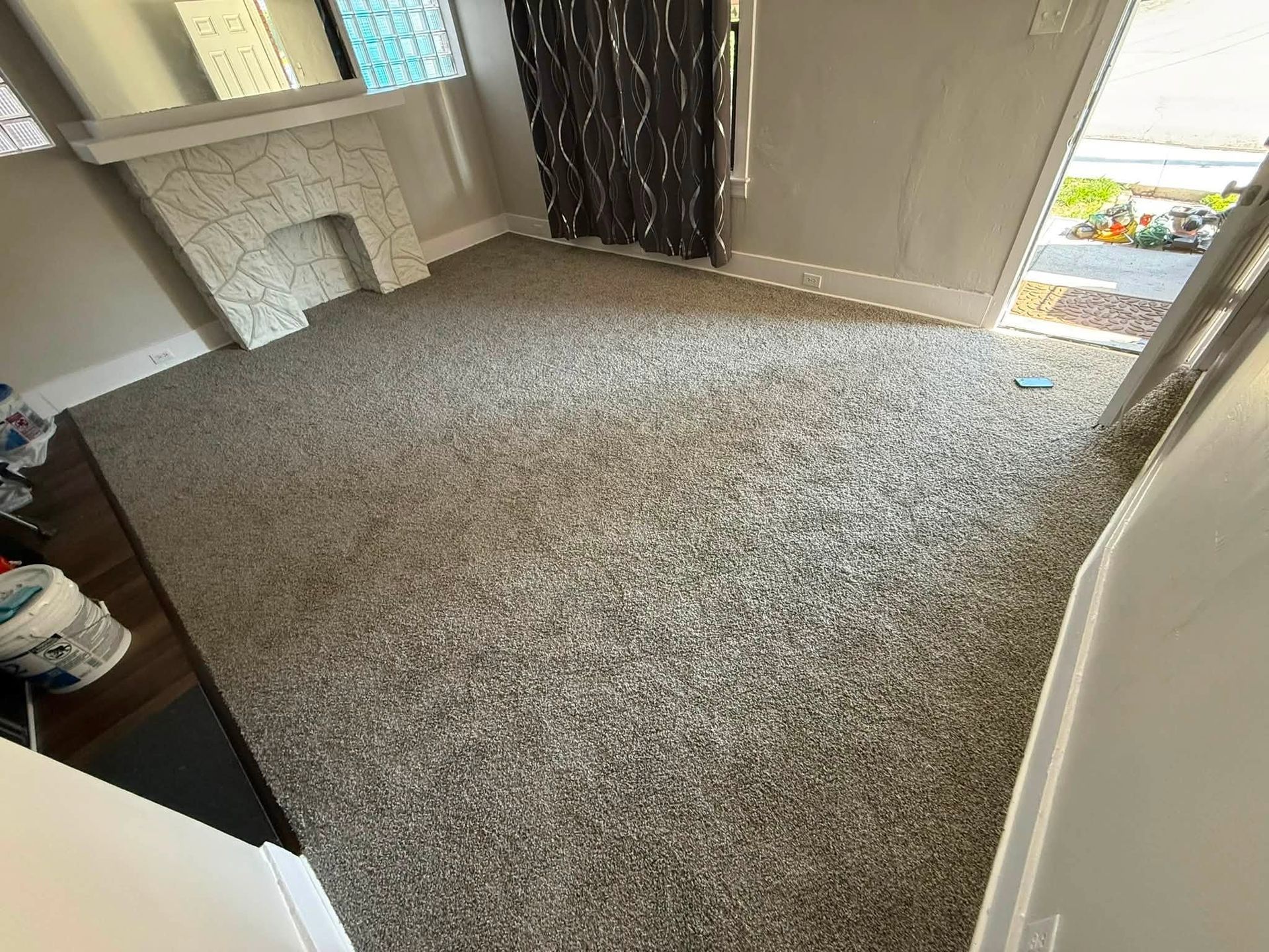 Empty carpeted living room with beige walls, a fireplace, dark curtains, and daylight through a glass door.