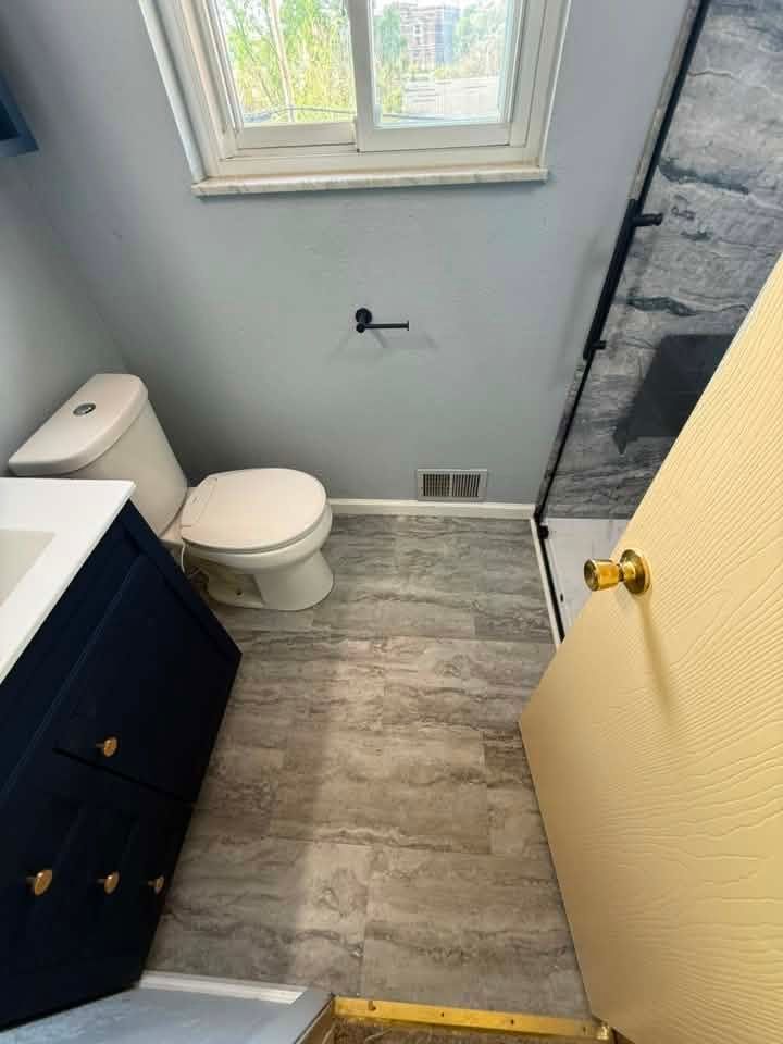 Small bathroom with toilet, vanity, window, and tiled floor; open door on right.