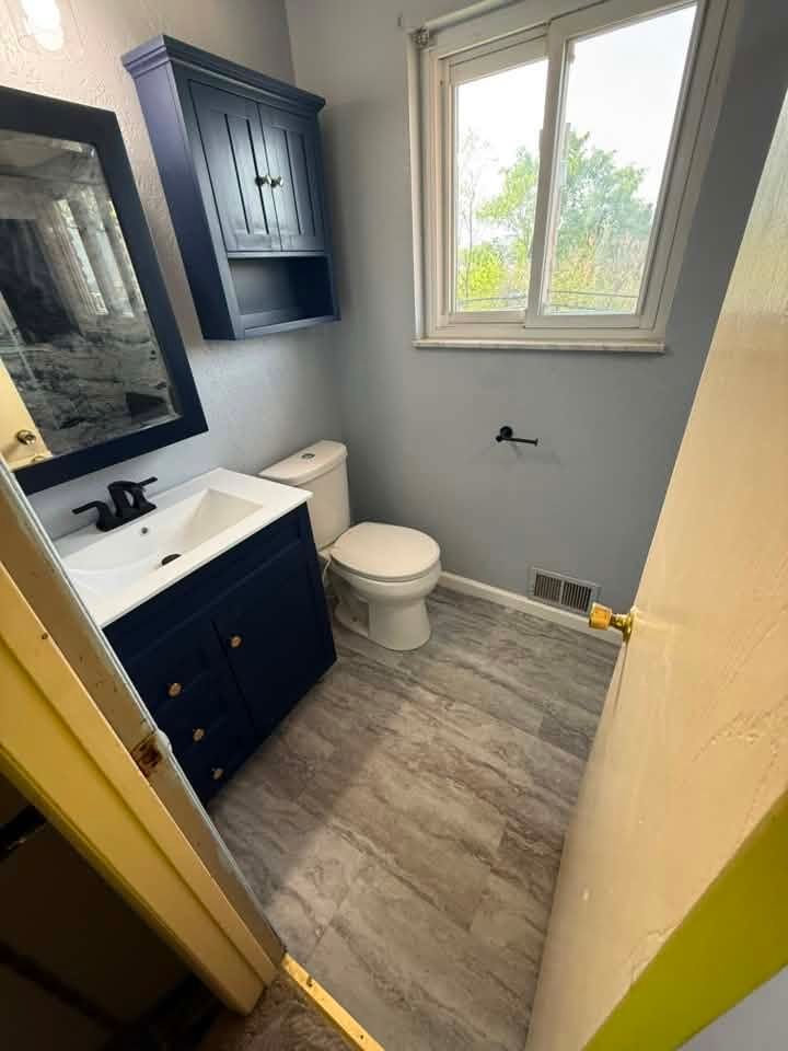 Small bathroom with white sink, toilet, blue vanity, gray flooring, and a window letting in daylight.