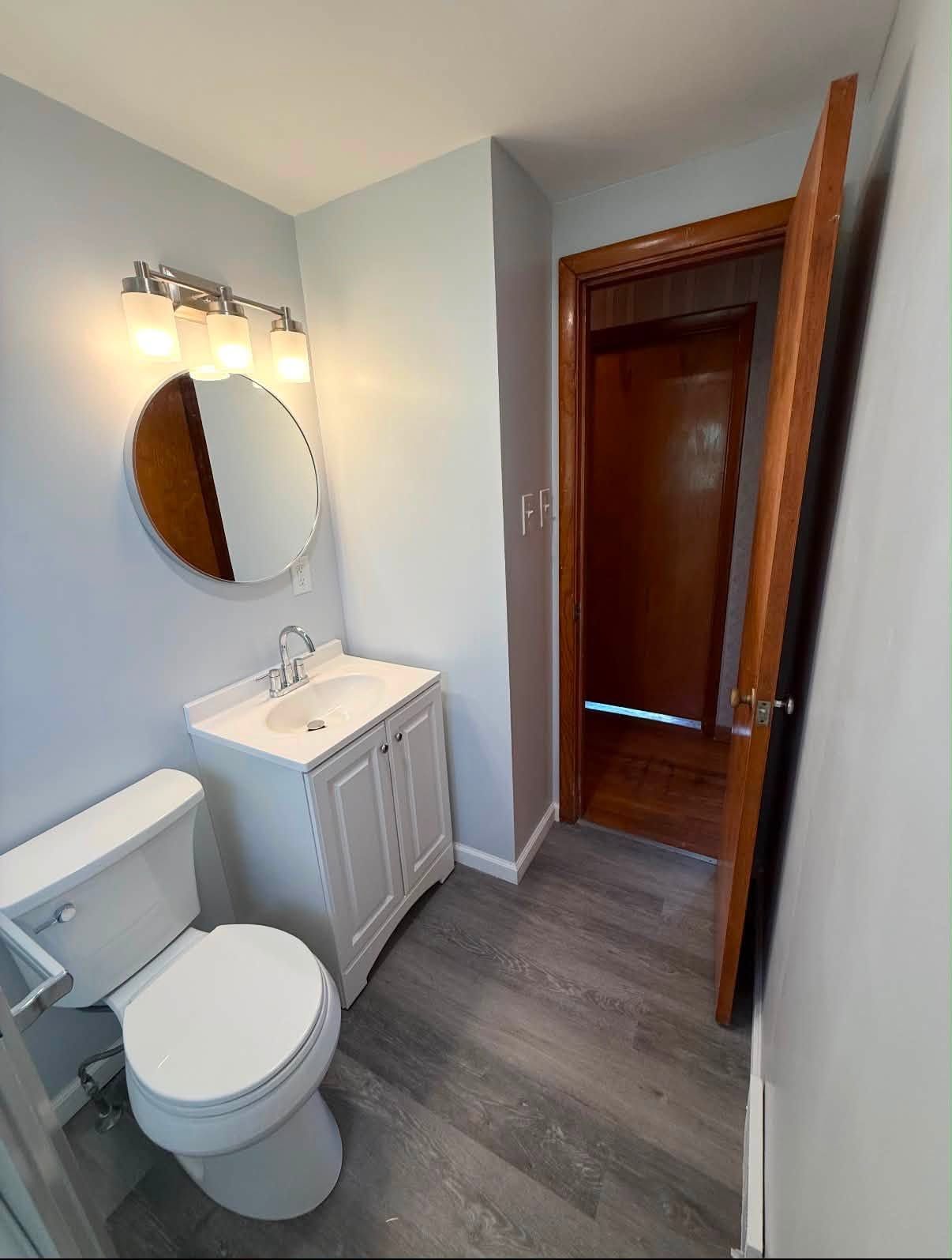 Small bathroom with white vanity, round mirror, toilet, gray floor, and open door to a hallway.