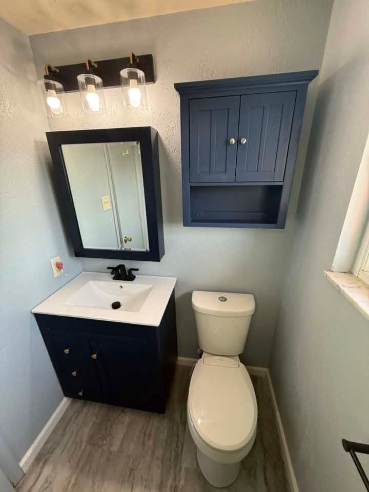 Small bathroom with white toilet, dark vanity and mirror, blue wall cabinet, and gray wood flooring.