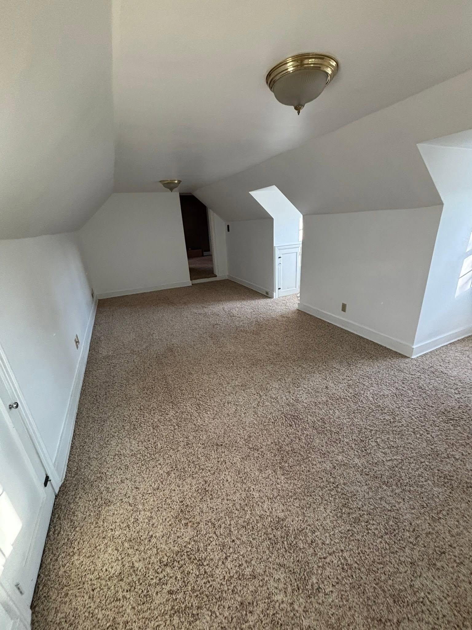 Empty carpeted attic room with sloped white ceilings and a brass ceiling light