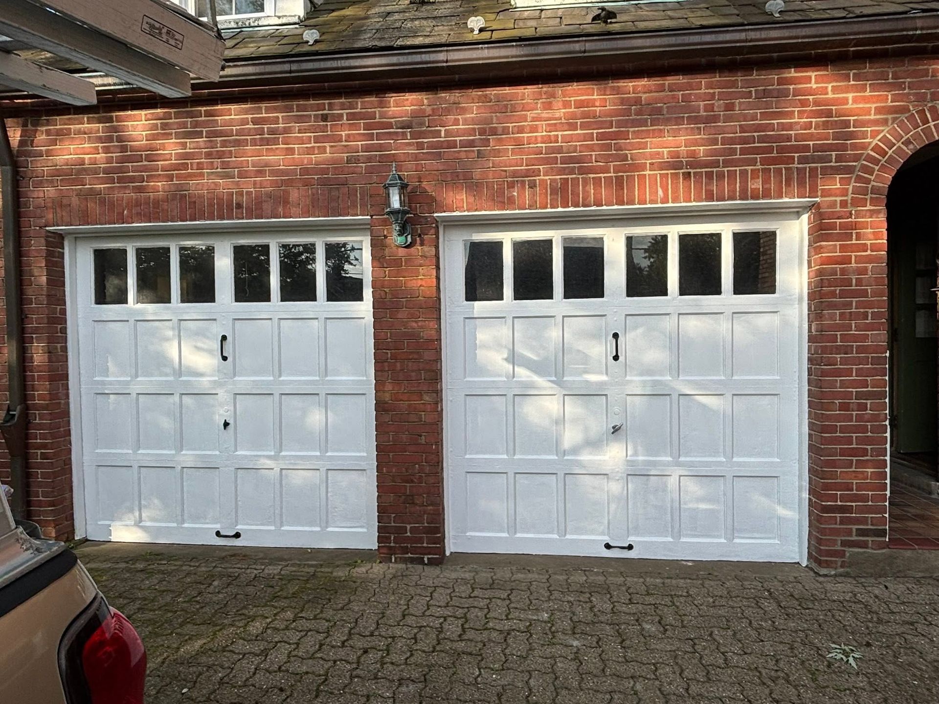 Two white garage doors set in a red brick wall, with a cobblestone driveway in front.