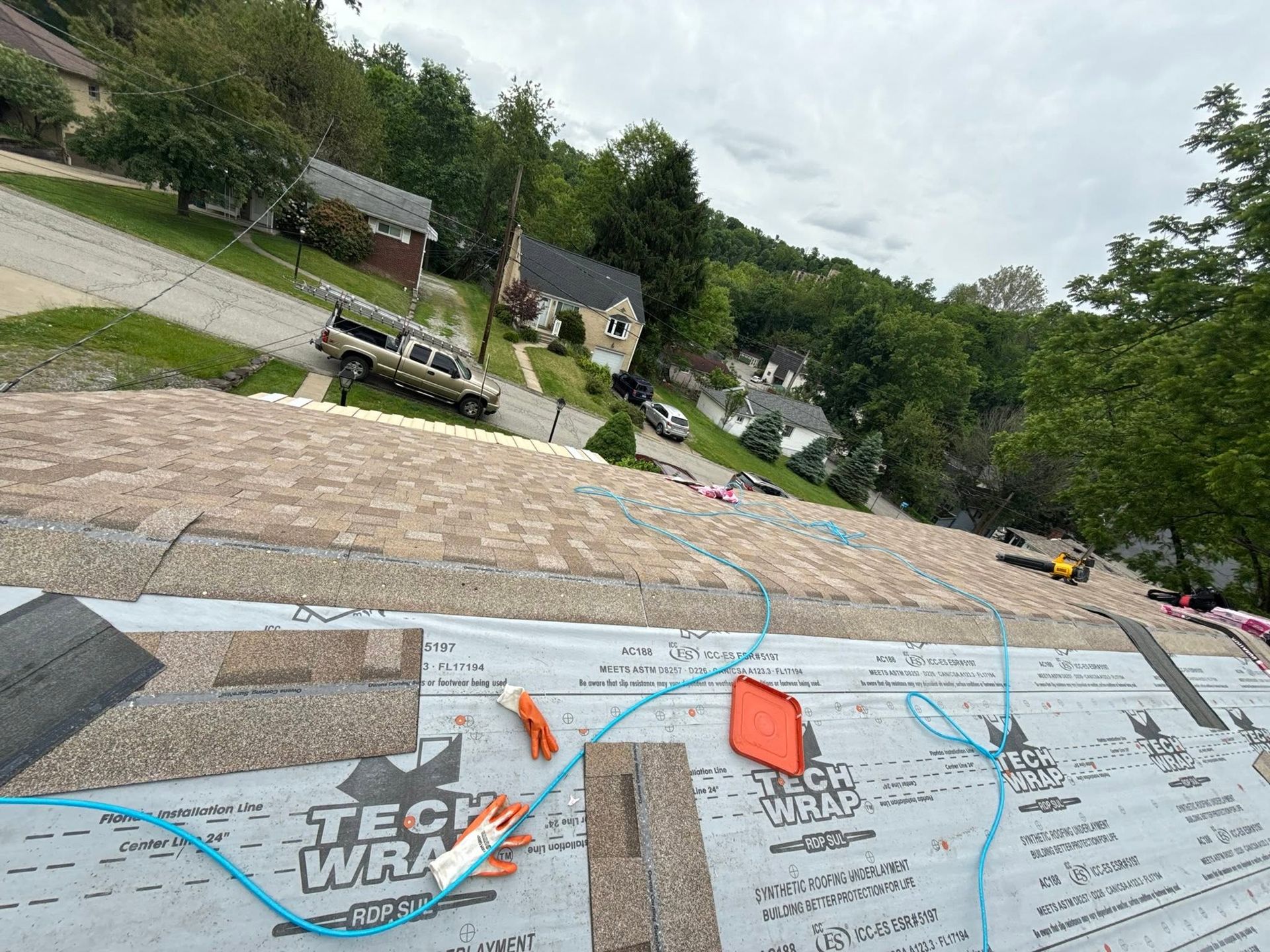 Roofer’s-eye view of a steep shingled roof with tools and an underlayment label, trees and houses below.