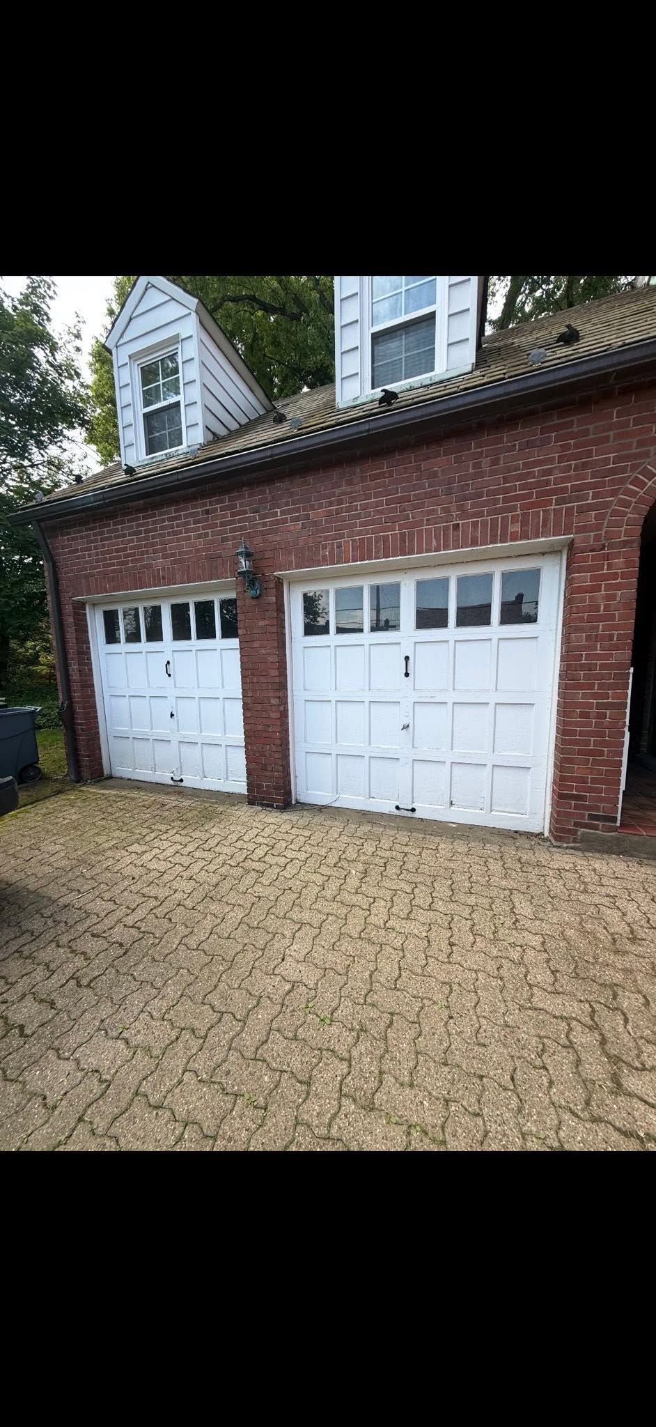 Red-brick two-car garage with white doors and gravel driveway, trees and apartment buildings behind.