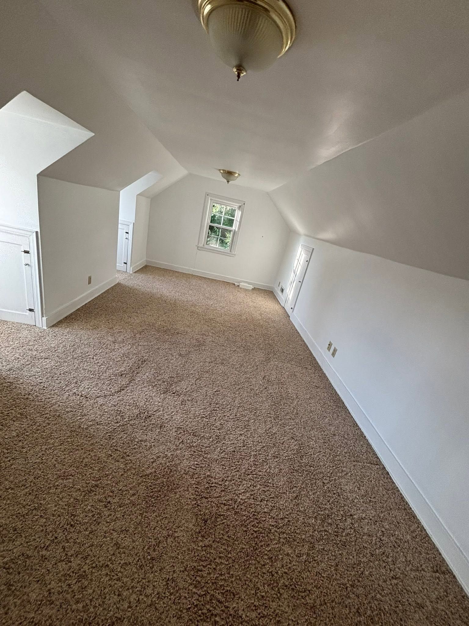 Empty beige-carpeted attic room with sloped white ceiling and small window end.
