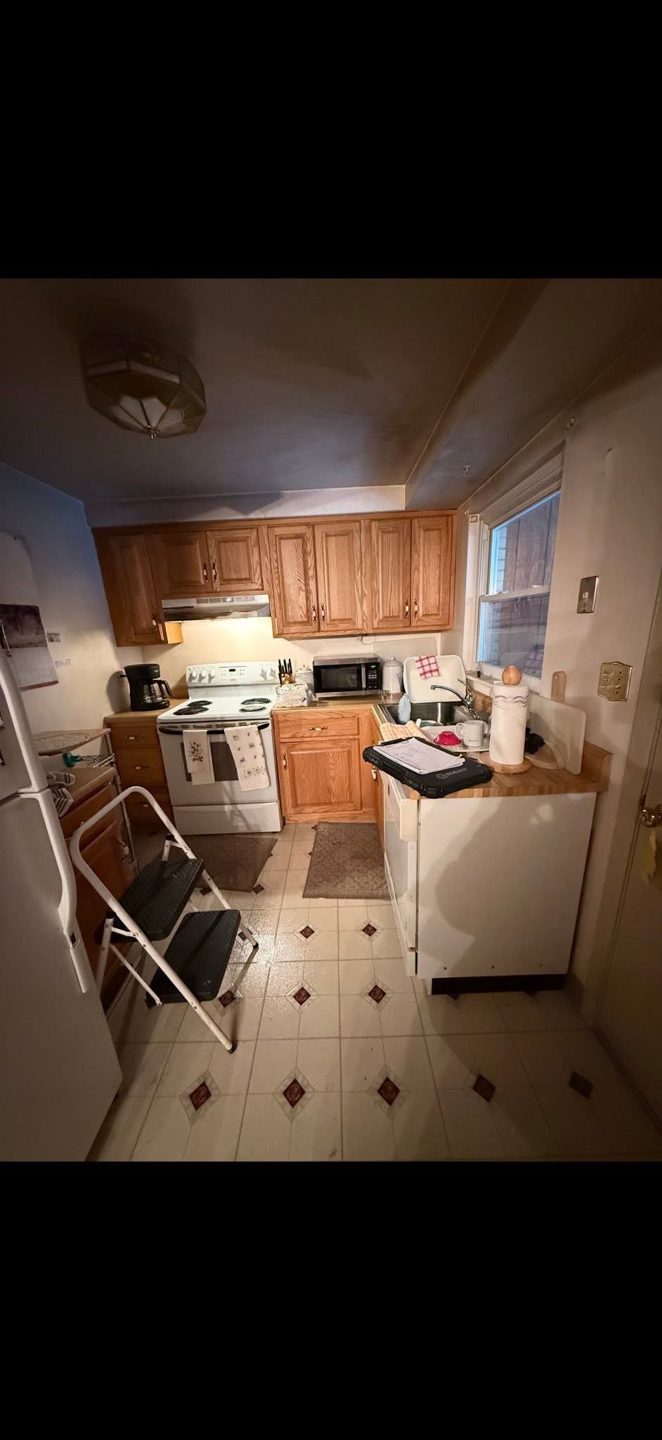 Small cluttered kitchen with wooden cabinets, stove, washing machine, and folding chairs on a tiled floor.