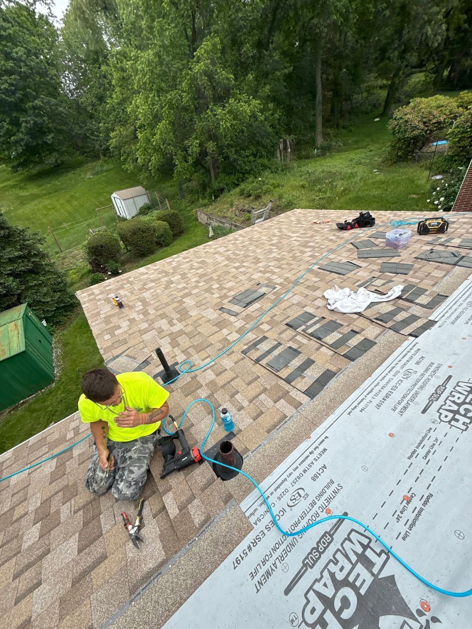 Worker on a rooftop installing shingles with tools and roofing materials nearby.
