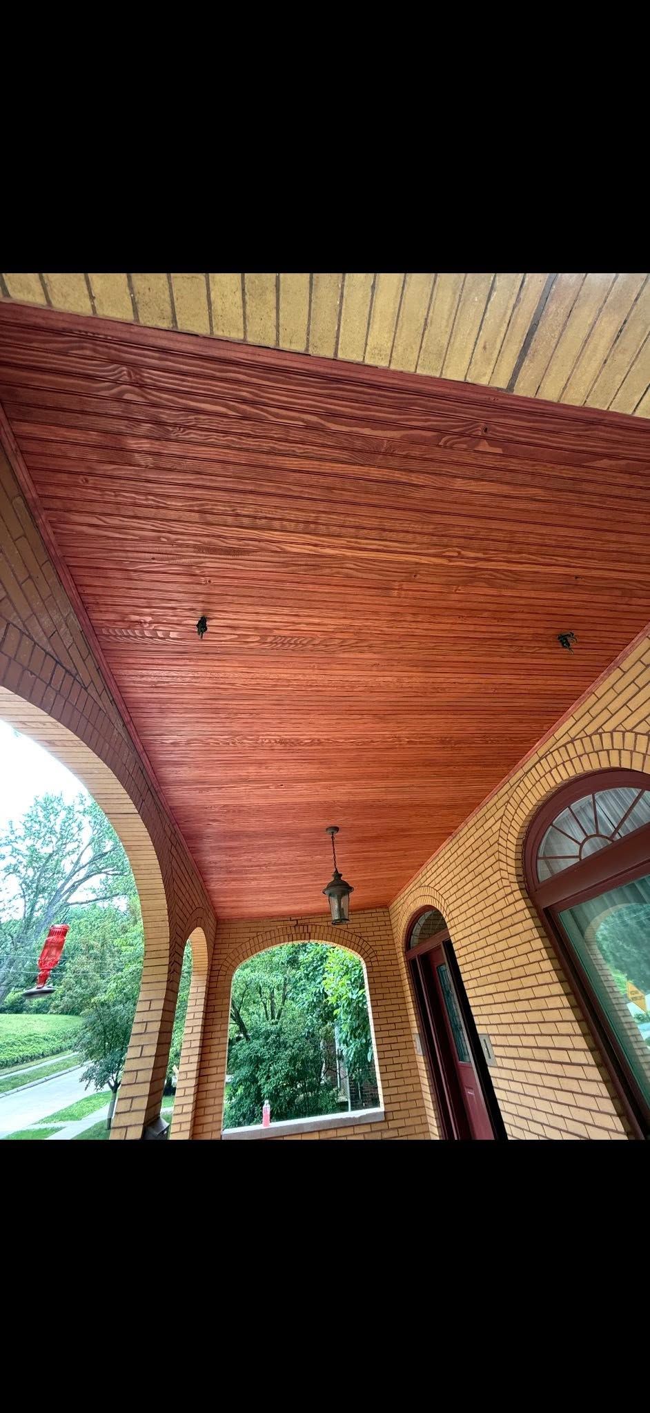 Brick-arched porch ceiling with wooden panels and hanging lights, framed by greenery outside the openings