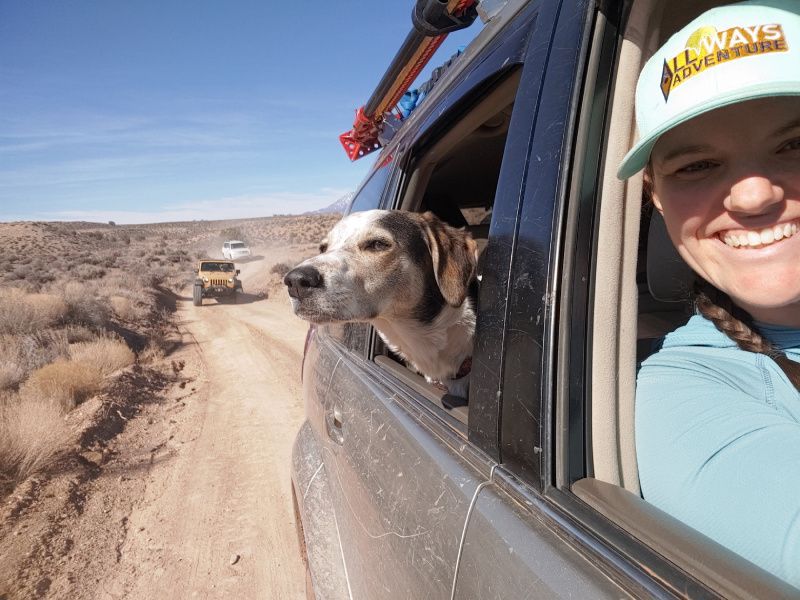 Woman smiles in car with dog looking out window inside the vehicle
