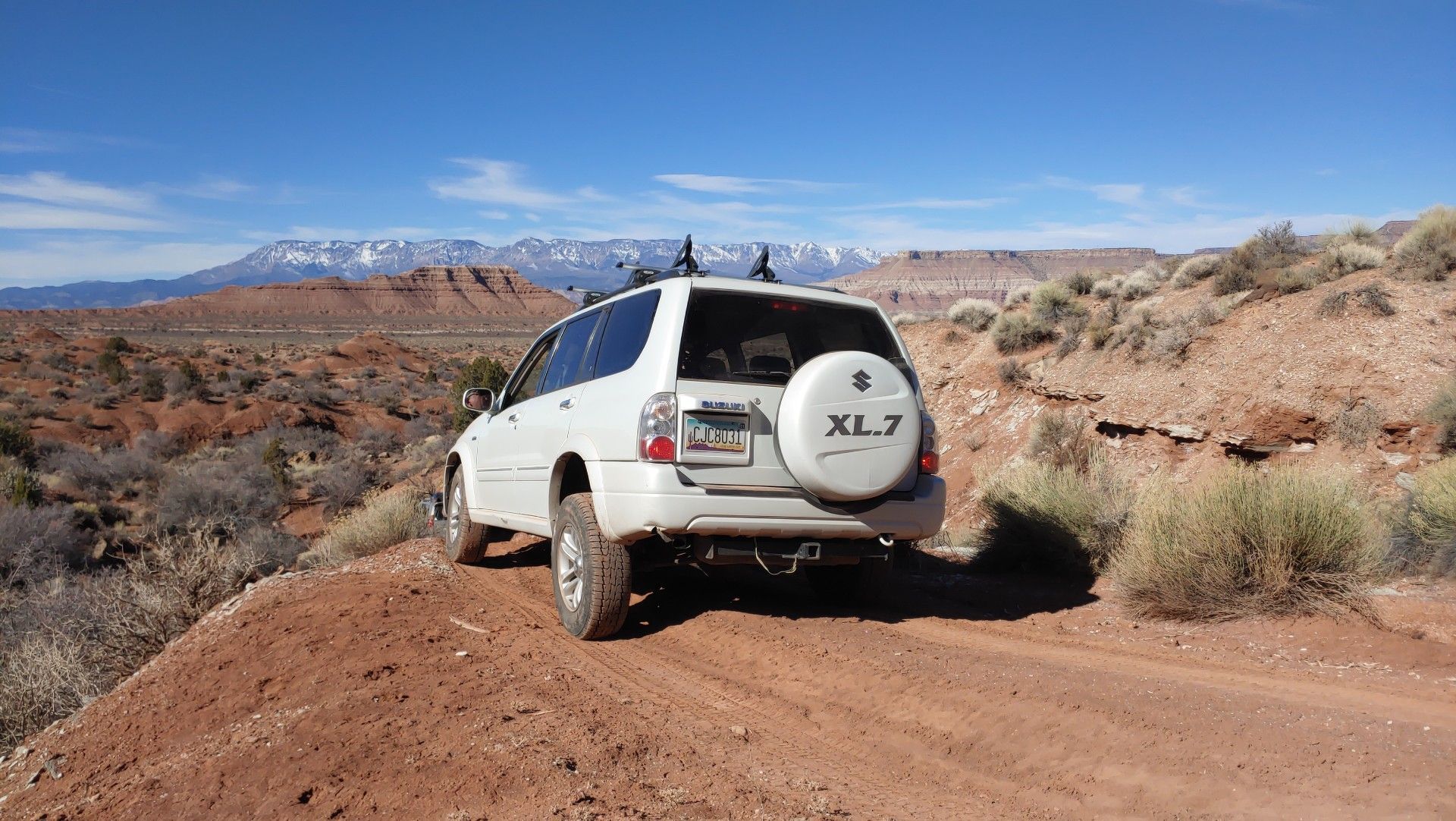White Suzuki XL7 SUV on a dirt road in a desert landscape with mountains in the background.