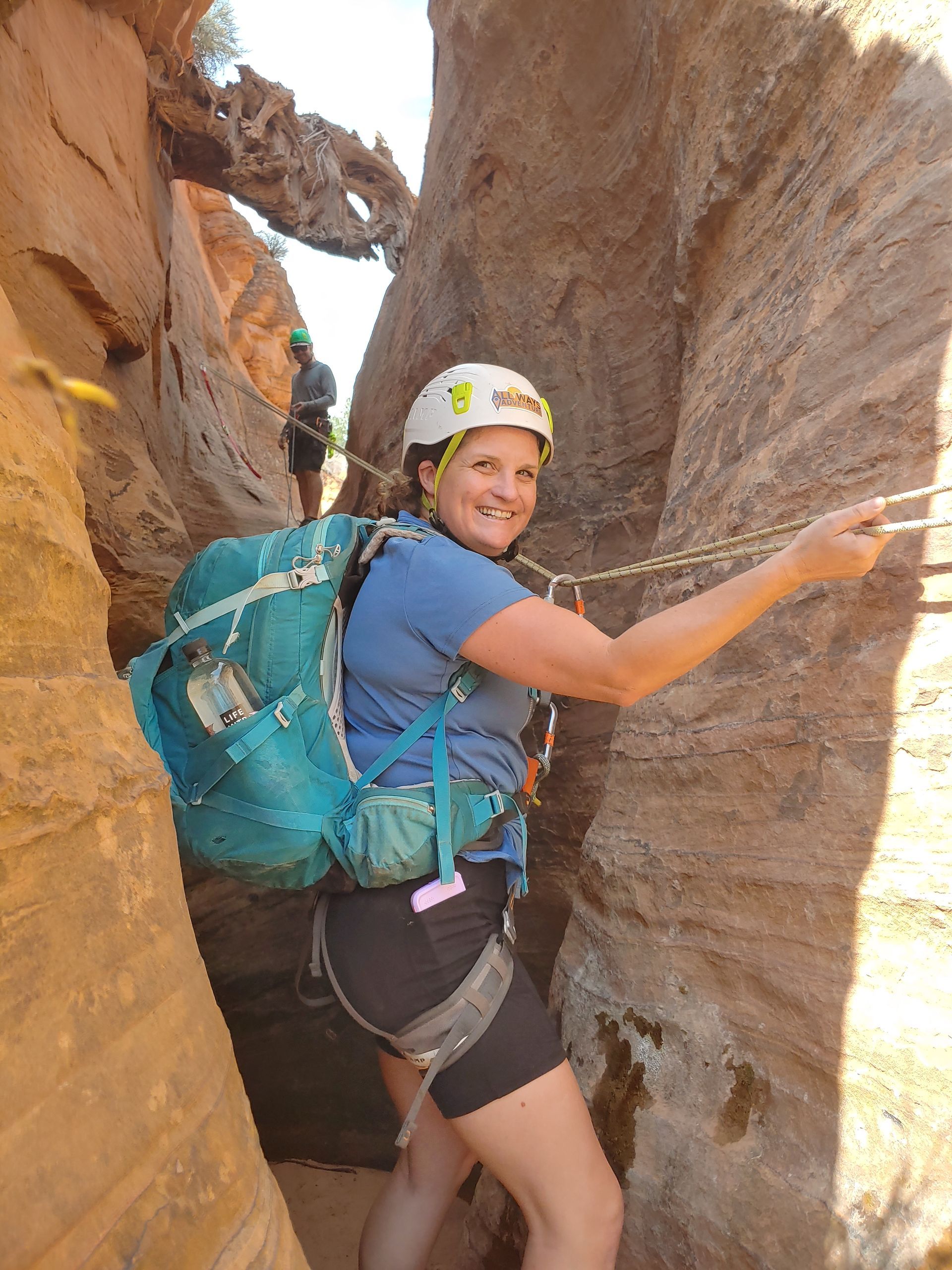 Woman in canyon, climbing with gear, smiling, using rope for support. Red rock walls.