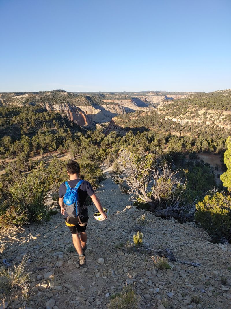A person with a backpack walks on a rocky cliff overlooking a canyon filled with trees under a clear blue sky.