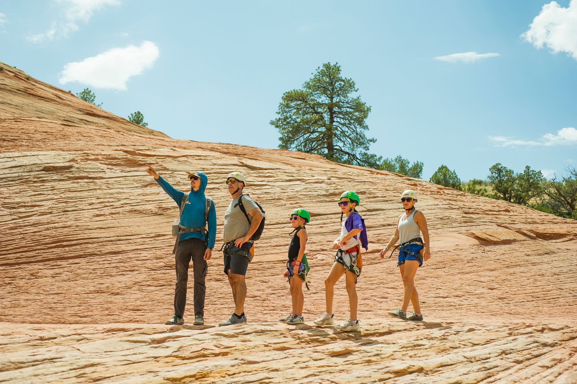 Five people, wearing helmets and harnesses, stand on a rocky hillside, one pointing, under a blue sky.