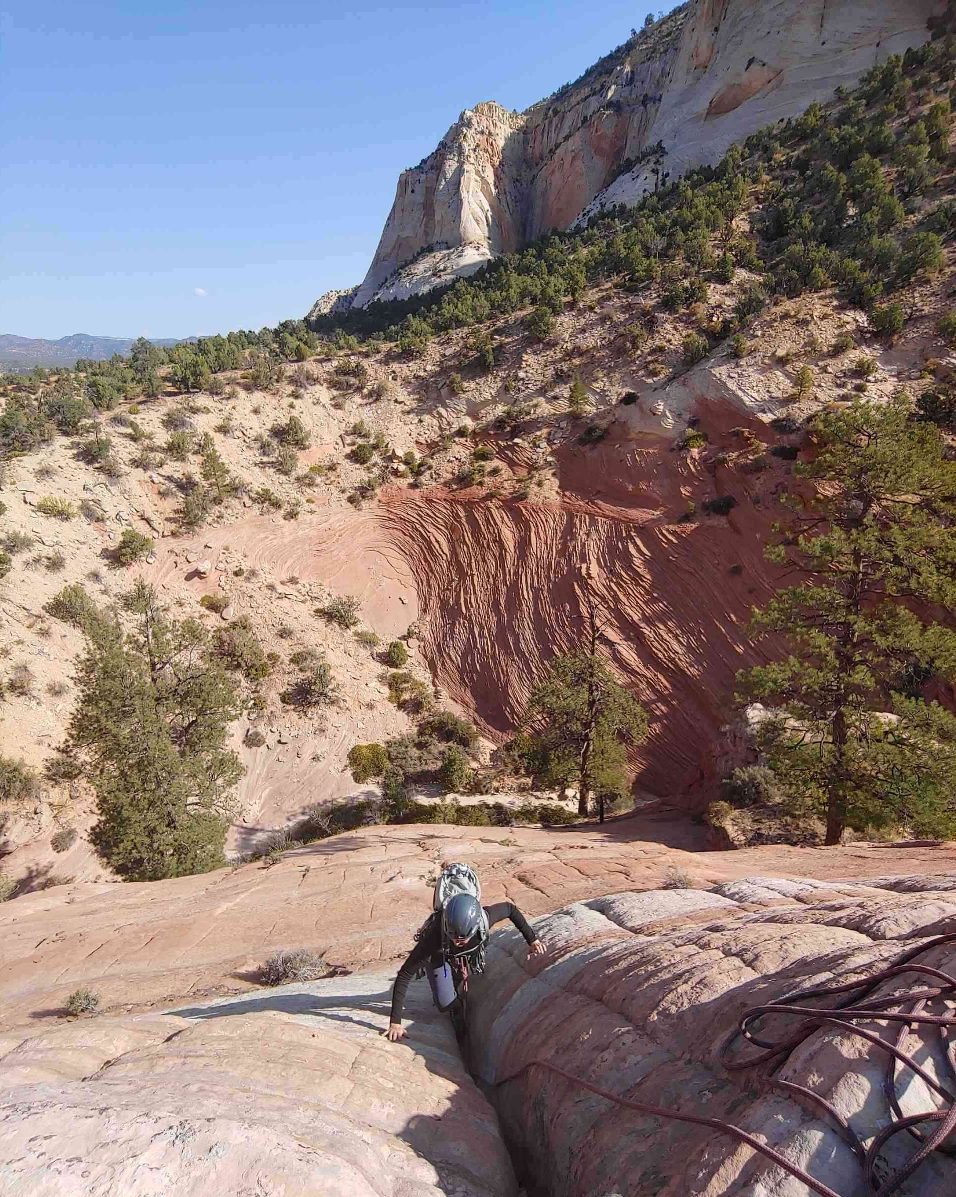 A person climbs a rocky hillside with gear, descending into a crevice, under a sunny sky.