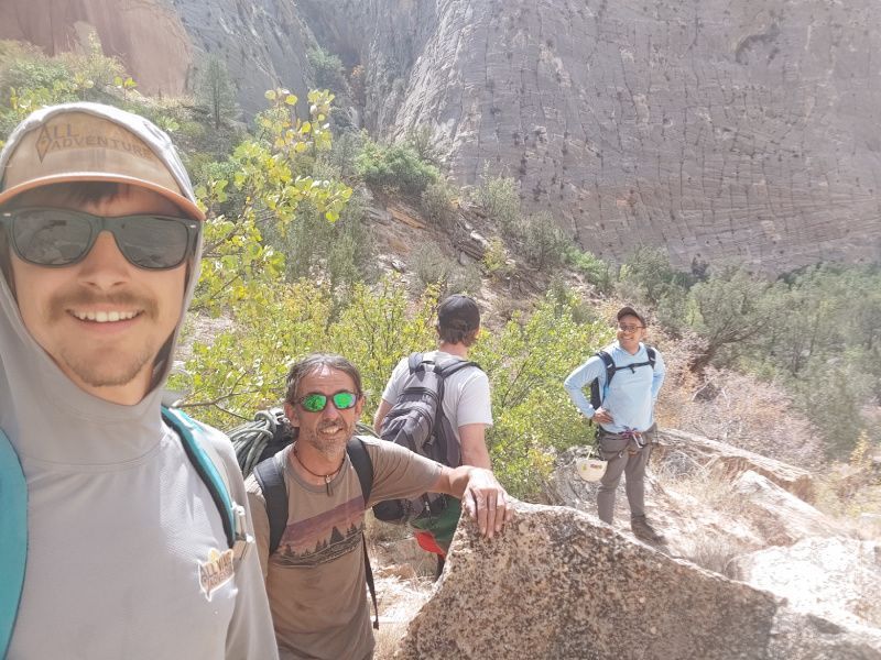Four hikers on a rocky path in a canyon. One takes a selfie; the others are hiking and looking at the view.