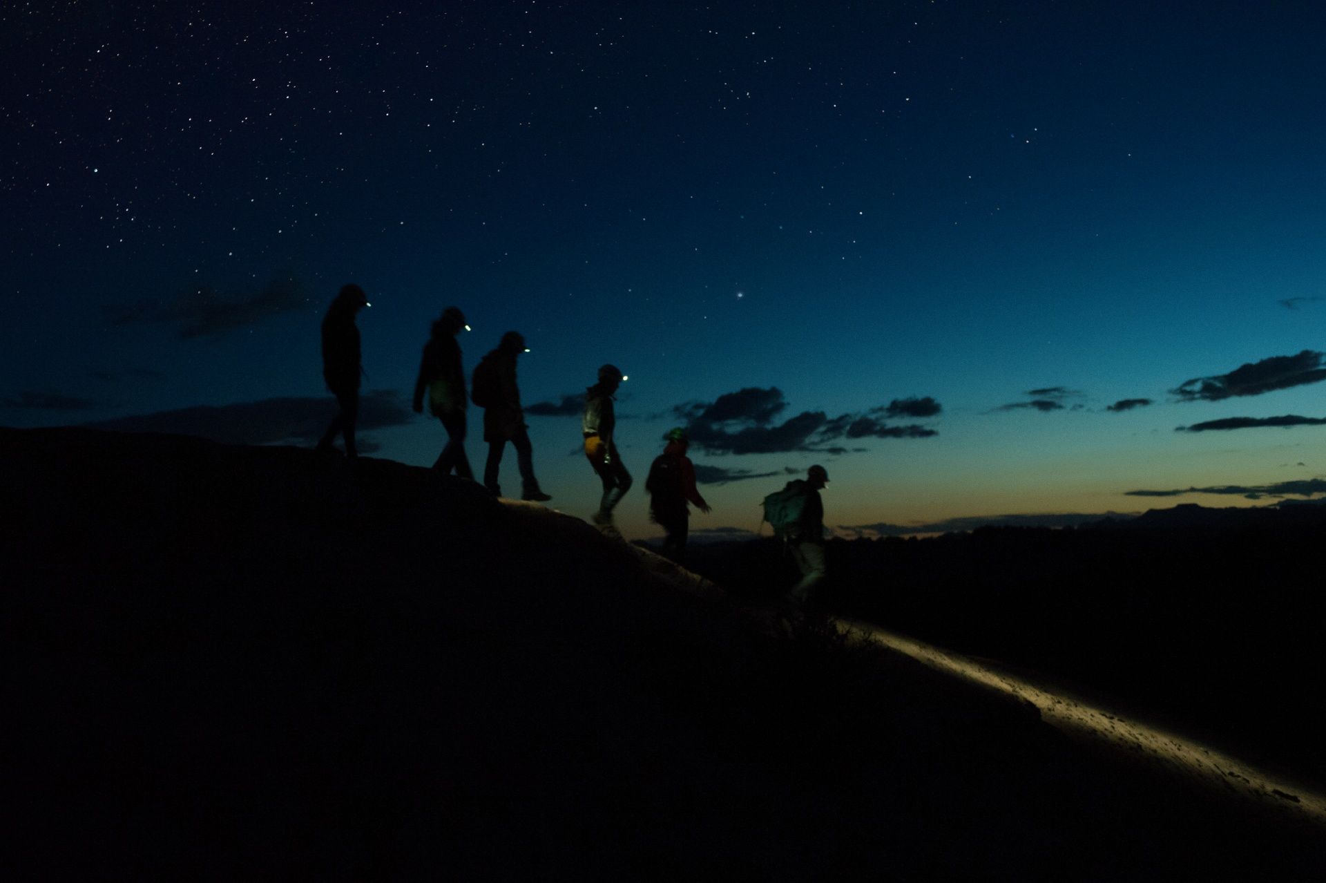Silhouetted hikers with headlamps walk across a ridge under a starry night sky.
