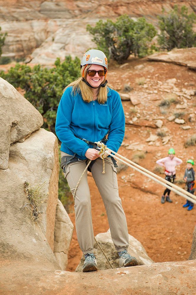 Woman in a helmet, blue jacket, and khaki pants rappels down a rock face; two children watch in the background.