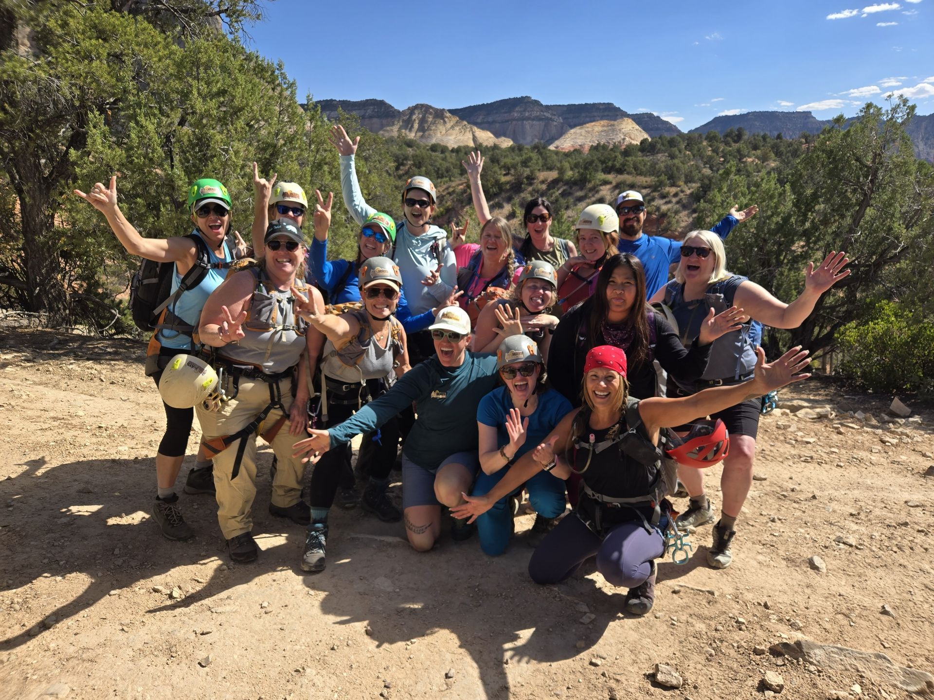 Group of hikers posing on a dusty trail with mountains and trees in the background, smiling and waving.
