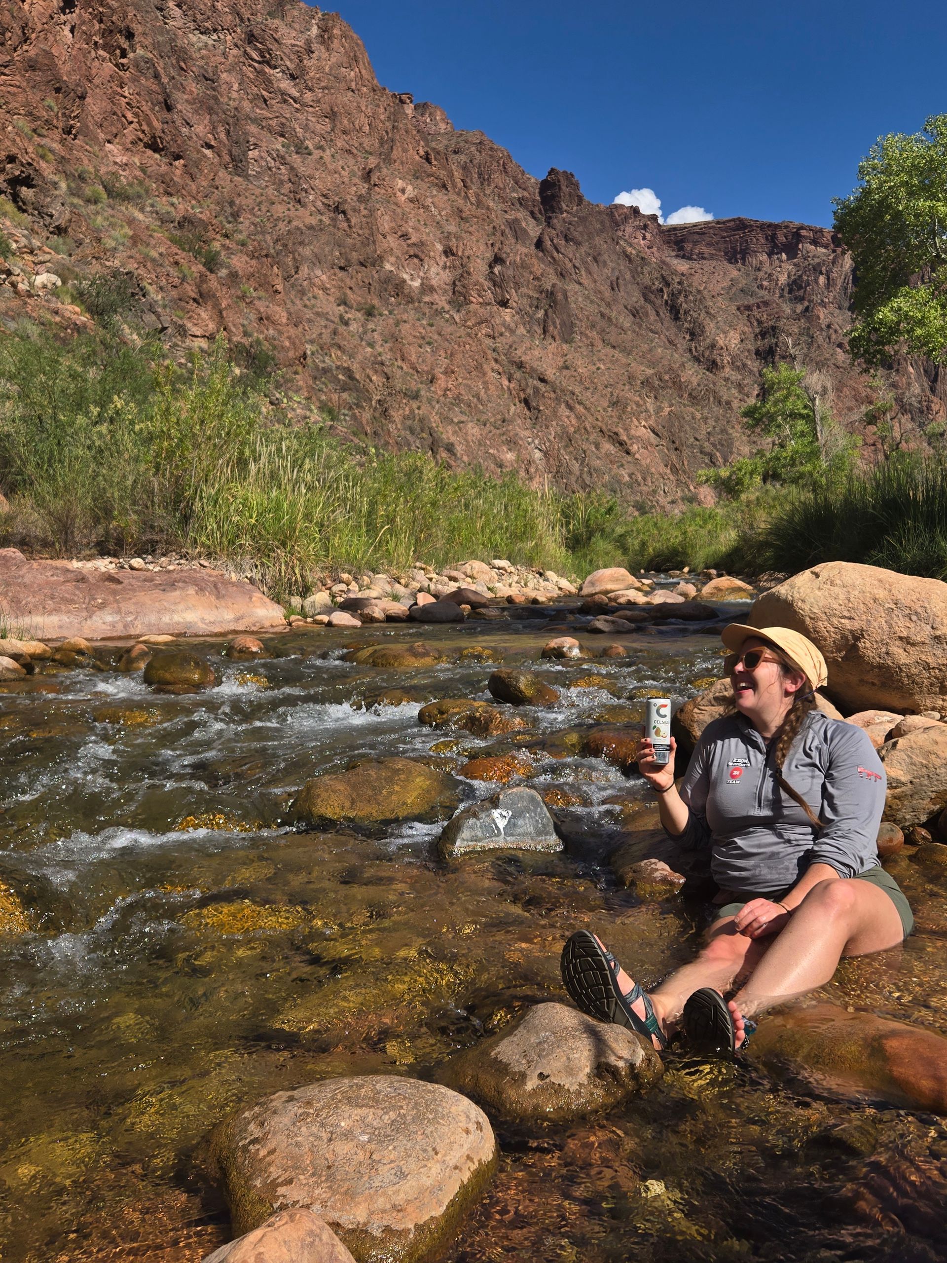 Woman smiles, holding drink while sitting in a rocky stream. She wears a hat, shirt, shorts.